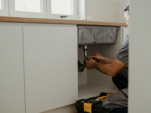 A technician's hands tightening a fitting under a kitchen sink, an open toolbox on the floor beside them.