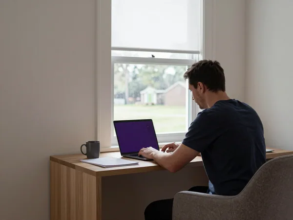 A man works alone at a home-office desk by a window, a laptop open beside a mug and a small stack of paperwork.